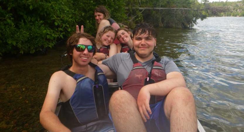 A group of teens wearing lifejackets sit in a canoe on calm water and smile for the photo. 